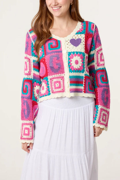 Woman in a colourful retro patchwork crochet cardigan featuring square patterns and a white skirt, smiling against a plain background.