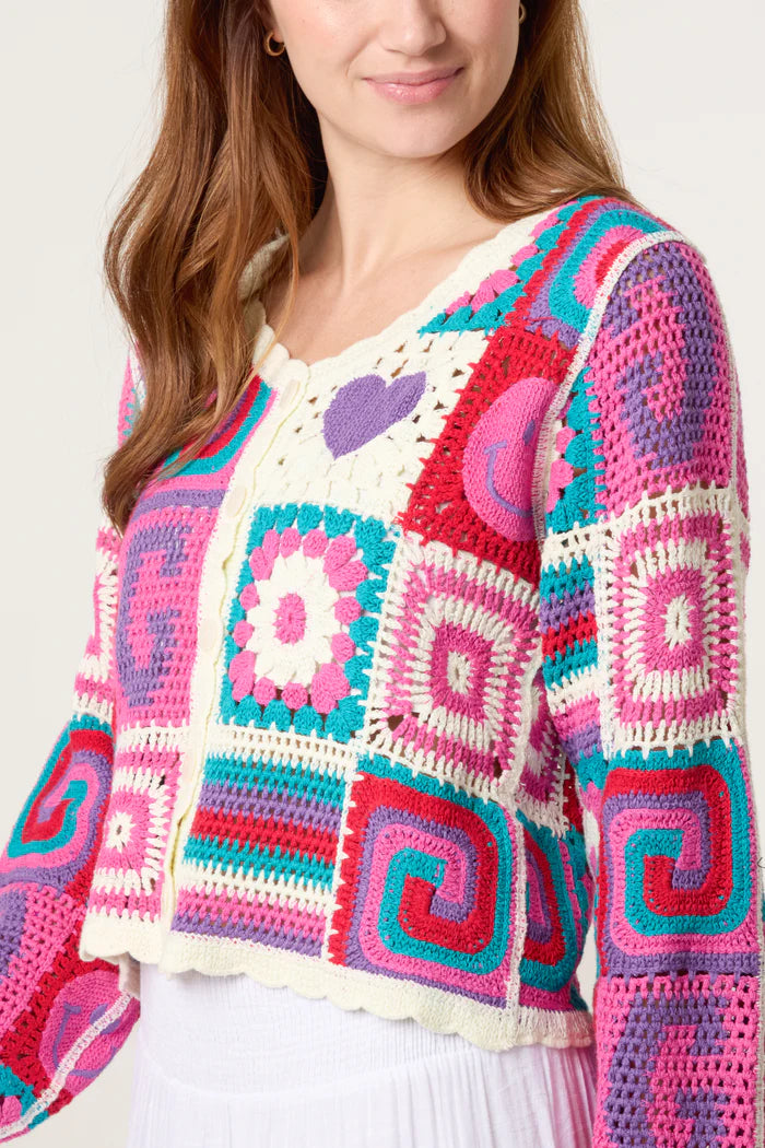 A woman wears a colourful retro patchwork crochet cardigan featuring square and swirl patterns in pink, purple, teal, and white, teamed with a white skirt.