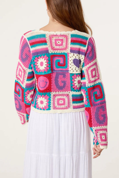 A person wearing a vibrant retro patchwork crochet cardigan featuring pink, purple, blue and white square patterns stands with their back to the camera.
