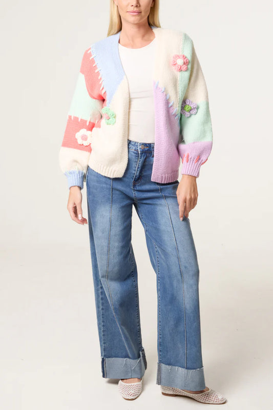 A woman wears a Stitch-inspired pastel patchwork cardigan with floral details, a white top, wide-leg cuffed jeans, and white trainers made from vegan materials, standing against a plain background.