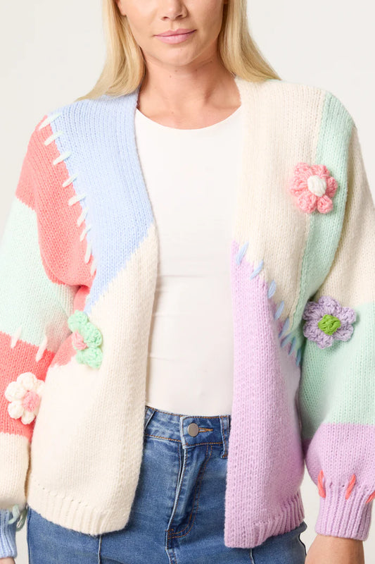 Woman wearing a pastel patchwork cardigan inspired by Stitch, featuring knitted flower details and made from sustainable vegan materials, layered over a white top and blue jeans, pictured from shoulders to hips.