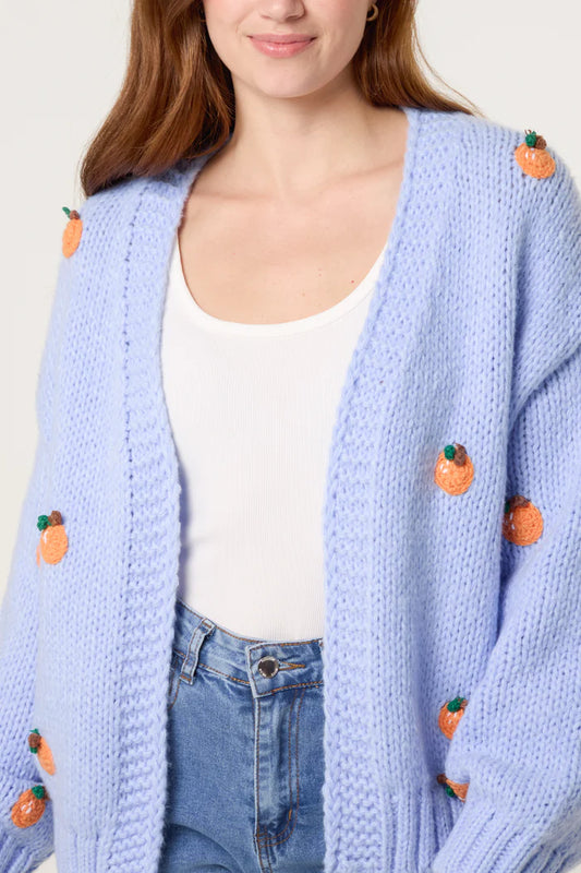 A woman is wearing a chunky blue cardigan decorated with small orange fruit appliqués, paired with a white vest top and blue jeans.