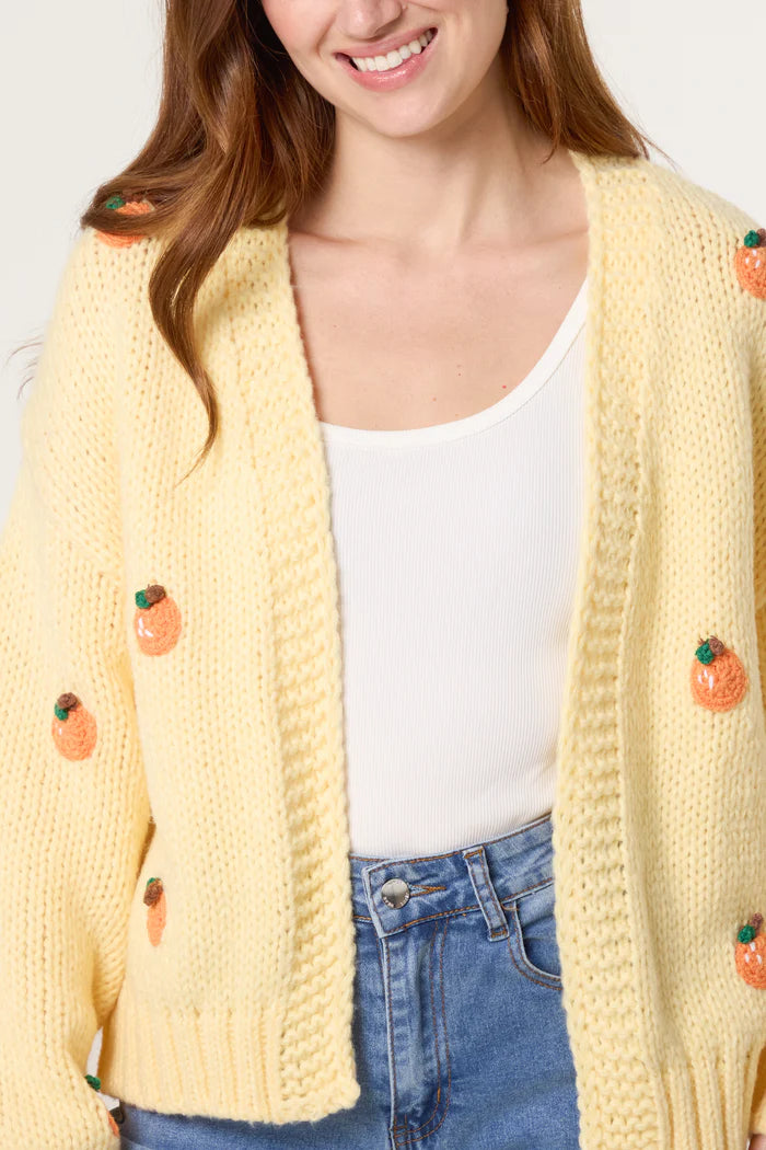 Woman wearing a pale yellow chunky knitted cardigan with orange fruit appliqués over a white top and blue jeans, smiling gently.
