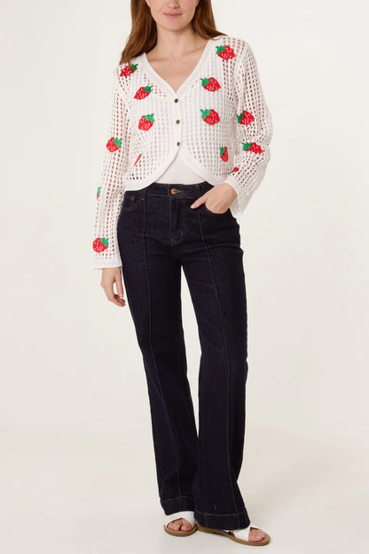 A woman is wearing a white strawberry crochet cardigan featuring red berry designs, a white top, dark high-waisted jeans, and white sandals, standing against a plain backdrop.