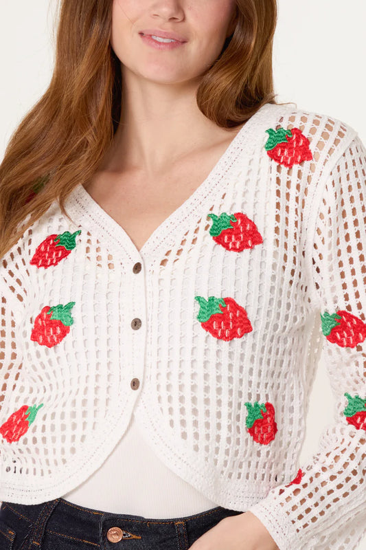 A woman wears a vintage strawberry crochet cardigan featuring red strawberry patterns over a white top and dark jeans.