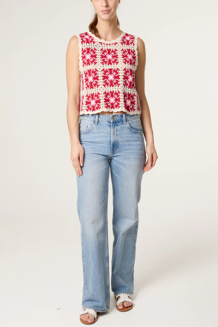 A woman stands wearing light blue jeans, white sandals, and a sleeveless red and white handmade vest top featuring a square crochet pattern.