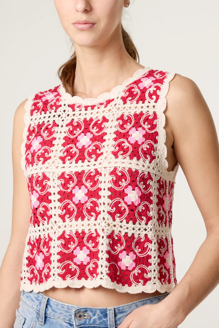 A woman wears a bohemian crochet top—a sleeveless red and cream floral design with scalloped edges—stylishly paired with light blue jeans.