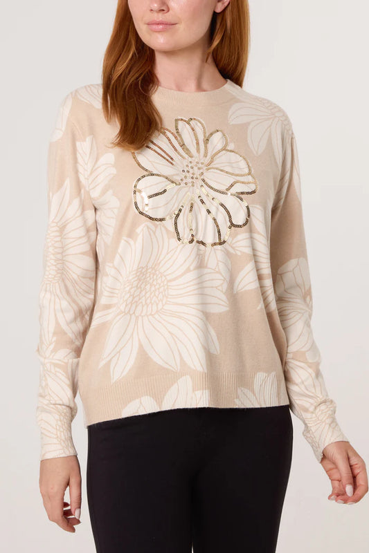 A woman is wearing a beige fine-knit crew neck jumper featuring large white embellished flowers and a gold sequin flower outline on the front, teamed with black trousers.