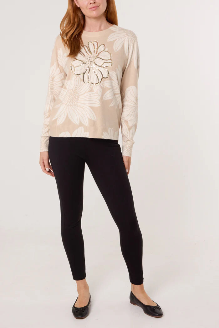 A woman stands against a plain background, dressed in a beige fine knit crew neck jumper with embellished flowers, teamed with black leggings and black ballet flats.