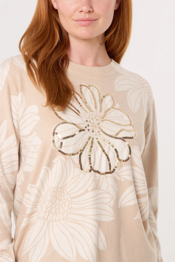 A woman wears a finely knitted crew neck jumper in beige, decorated with large embellished flowers and a central floral motif outlined with gold and black sequins.
