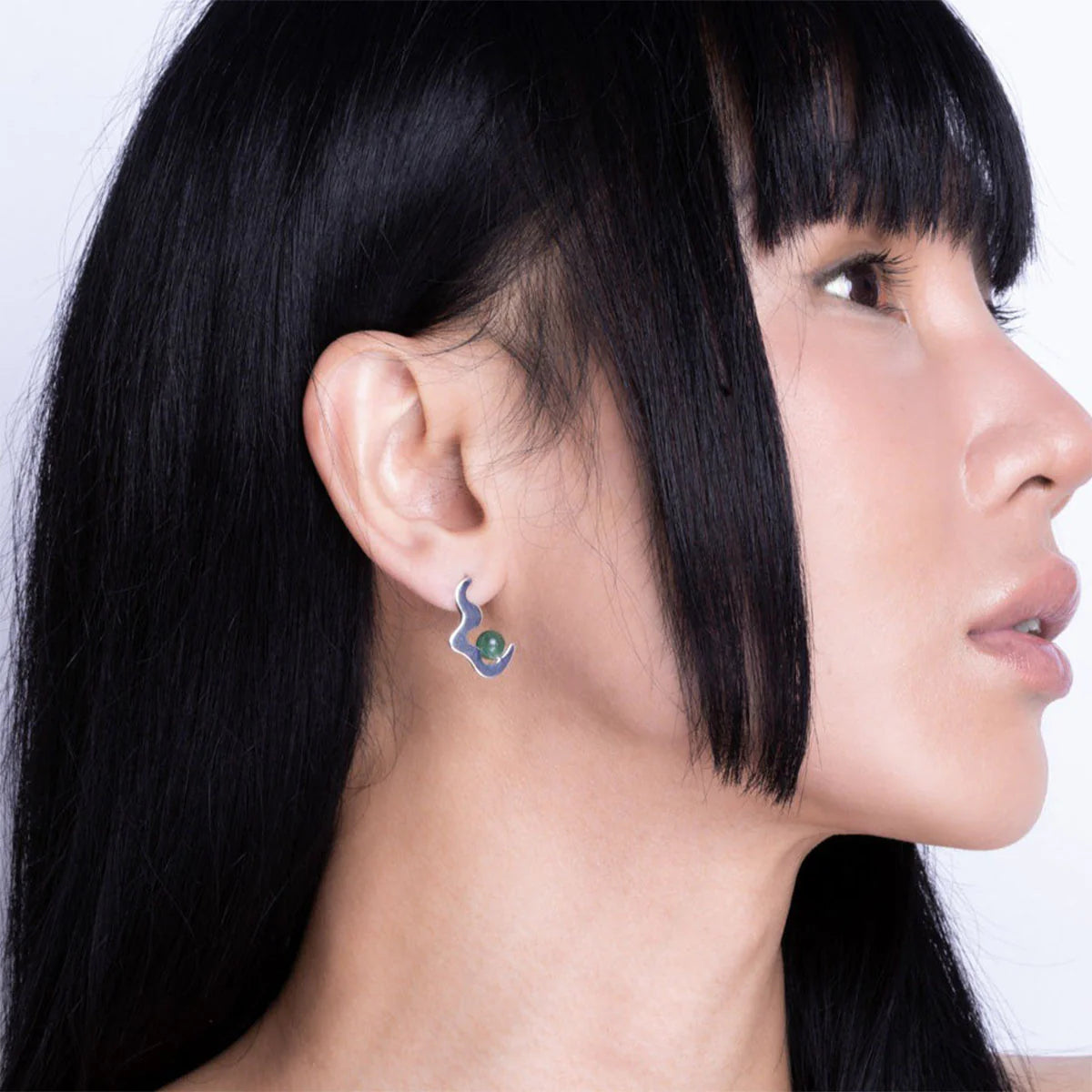 Individual with long black hair wearing a silver, snake-shaped aventurine earring featuring a green gemstone, shown in profile against a plain backdrop.