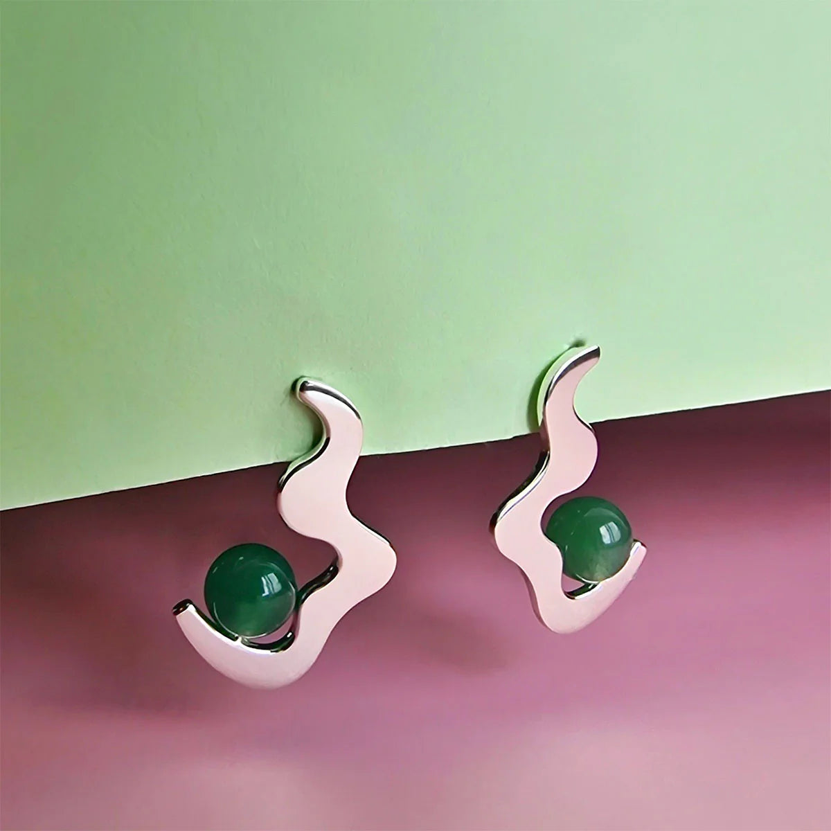 A pair of abstract wave drop studs featuring wavy silver shapes and green aventurine beads, set against a pastel green and pink background.