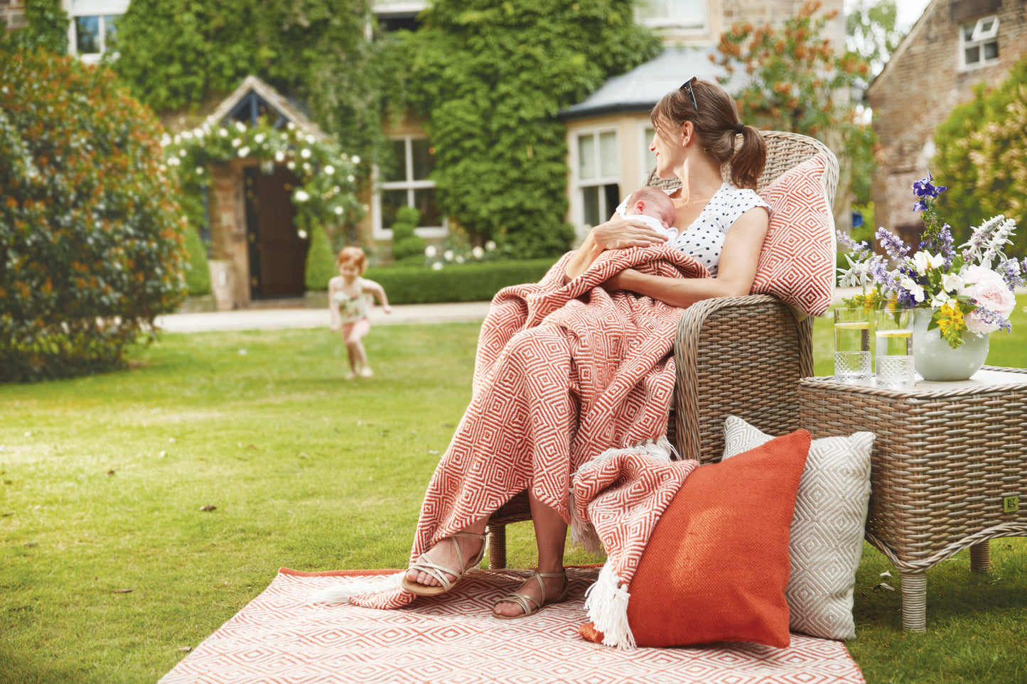 A woman sits on a wicker chair outdoors, wrapped in an eco-friendly throw made from recycled plastic bottles, holding a baby as a child runs towards her, with a house in the background.