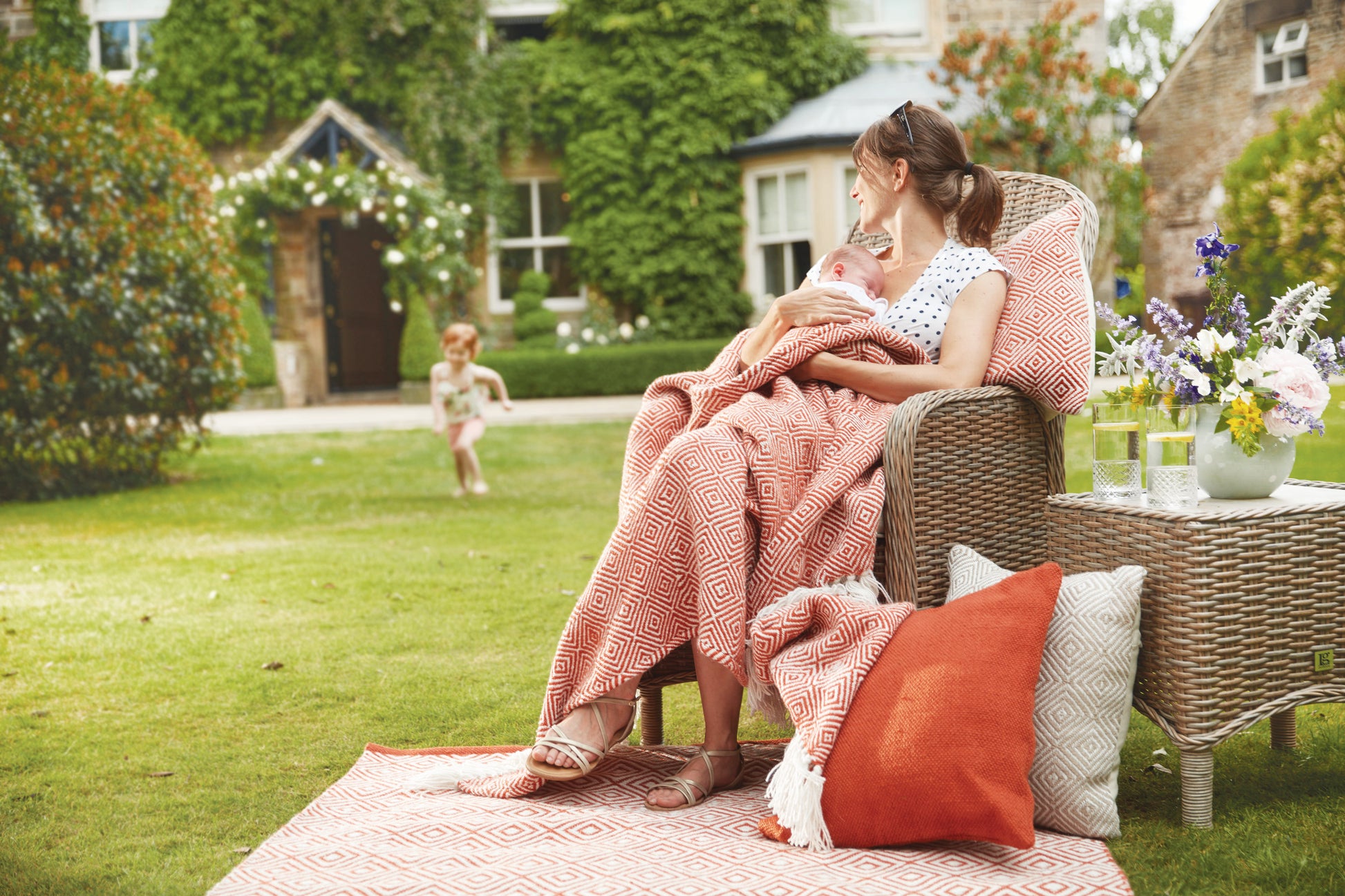 A woman sits on a wicker chair outdoors, wrapped in an eco-friendly throw made from recycled plastic bottles, holding a baby as a child runs towards her, with a house in the background.