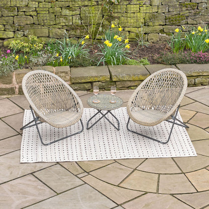 Two rattan chairs and a small round table with two glasses of wine create an inviting garden furniture set on a patterned rug along the stone patio, with blooming flowers in the background. Revised in British English: Two rattan chairs and a small round table with two glasses of wine form an inviting garden furniture set on a patterned rug across the stone terrace, with blooming flowers in the background.