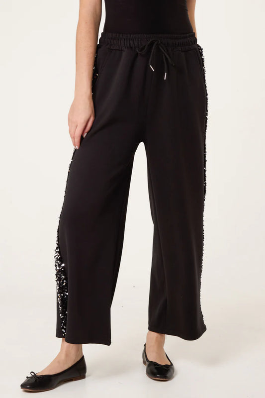 Individual dressed in sequin velvet joggers, black ballet flats, and a black top, highlighting chic Italian loungewear style against a simple light backdrop.