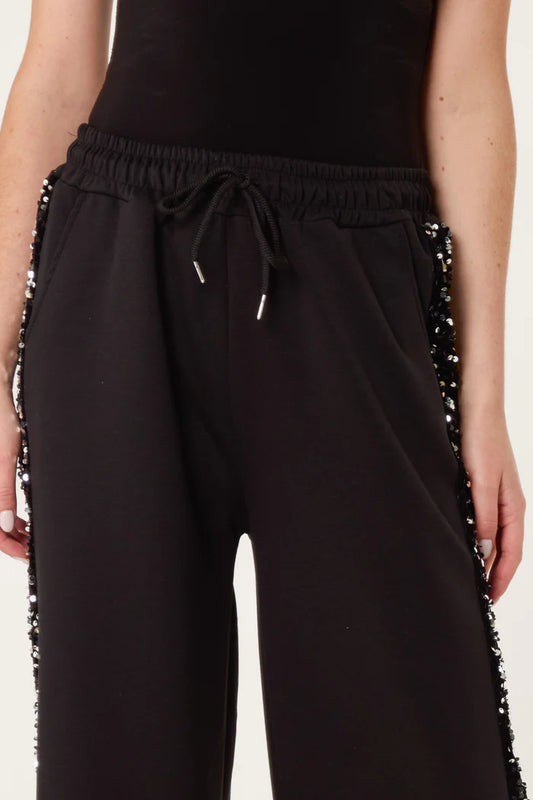 Person wearing black sequin velvet joggers with embellishments along the sides, paired with a black top for a stylish athleisure look.