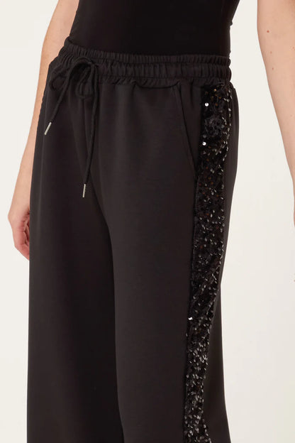 A person wearing black sequin velvet joggers with a drawstring waist and sequin detailing along the side seam, paired with a black top for a stylish athleisure look.