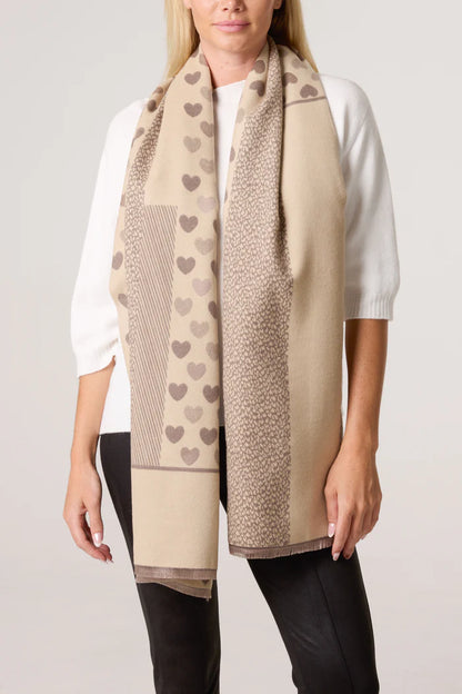 A woman in a white top and black trousers models a vegan-friendly, reversible scarf adorned with heart and geometric patterns in assorted shades of brown.