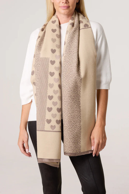 A woman in a white top and black trousers models a vegan-friendly, reversible scarf adorned with heart and geometric patterns in assorted shades of brown.
