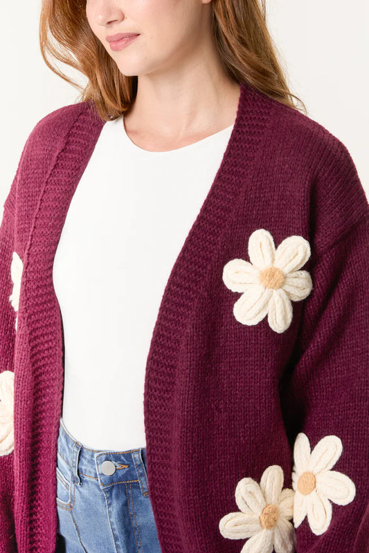 Woman wearing a hand-knitted cardigan adorned with large white flower appliqués over a white blouse and blue jeans, highlighting sustainable fashion.