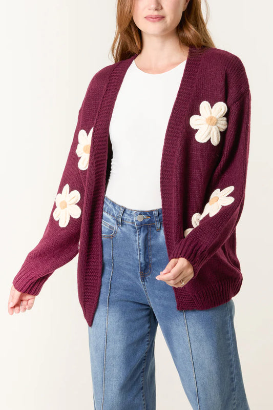 A woman wears a hand-knitted maroon cardigan with white daisy appliqués over a white top and blue jeans, highlighting sustainable fashion.