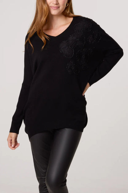 A woman wearing a black V-neck jumper with black floral embellishments on the shoulder and black leather trousers stands with one hand on her hip.