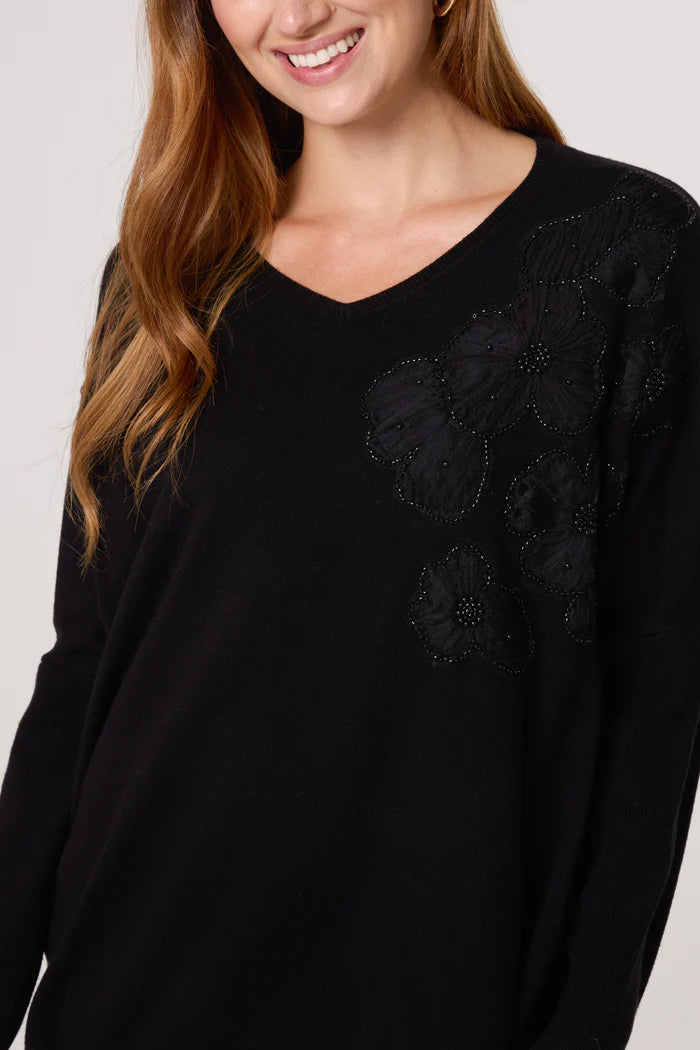 Woman wearing a black V-neck jumper featuring floral embellishments and beaded detailing on the shoulder, smiling with long light brown hair.