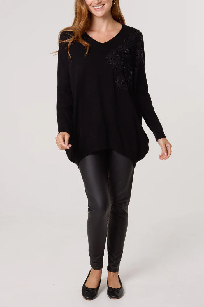 A woman is dressed in a black V-neck jumper adorned with black floral embellishments, teamed with black leather leggings and flat shoes, standing against a plain light-coloured background.
