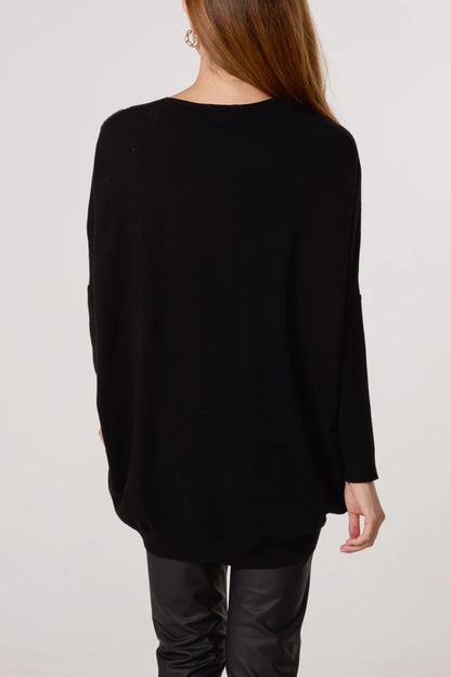 A person with long brown hair is seen from behind, wearing a loose-fitting V-neck jumper and black trousers, set against a plain light background.