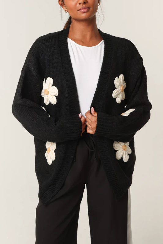 A person in a hand-knitted black cardigan adorned with large white daisy flower appliqués, worn over a white shirt and black trousers, highlighting sustainable style.