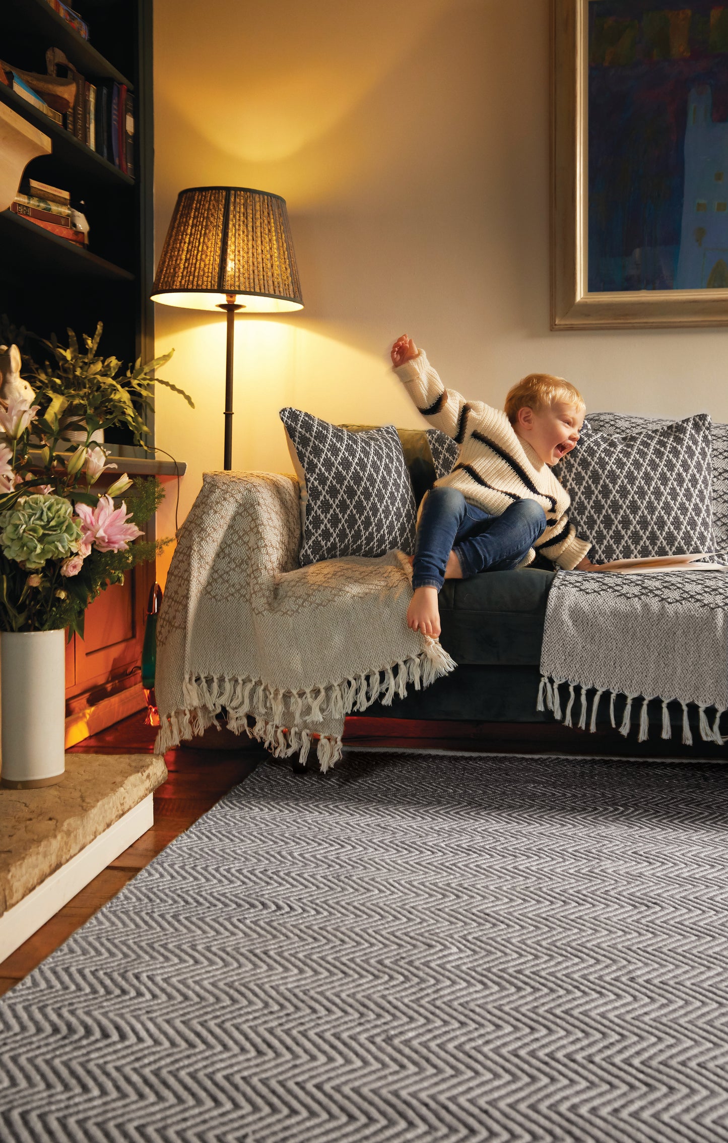 A child in a striped jumper and jeans stretches on a grey sofa, surrounded by patterned cushions and a fringed throw. Nearby, a lamp glows beside a vase of flowers on the shelf, while below lies an eco-friendly rug made from recycled plastic bottles.