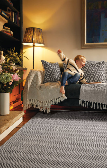 A child in a striped jumper and jeans stretches on a grey sofa, surrounded by patterned cushions and a fringed throw. Nearby, a lamp glows beside a vase of flowers on the shelf, while below lies an eco-friendly rug made from recycled plastic bottles.