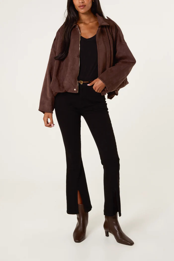 A woman in a chocolate suedette jacket, black top, black trousers with front slits, and brown ankle boots stands against a plain light background.