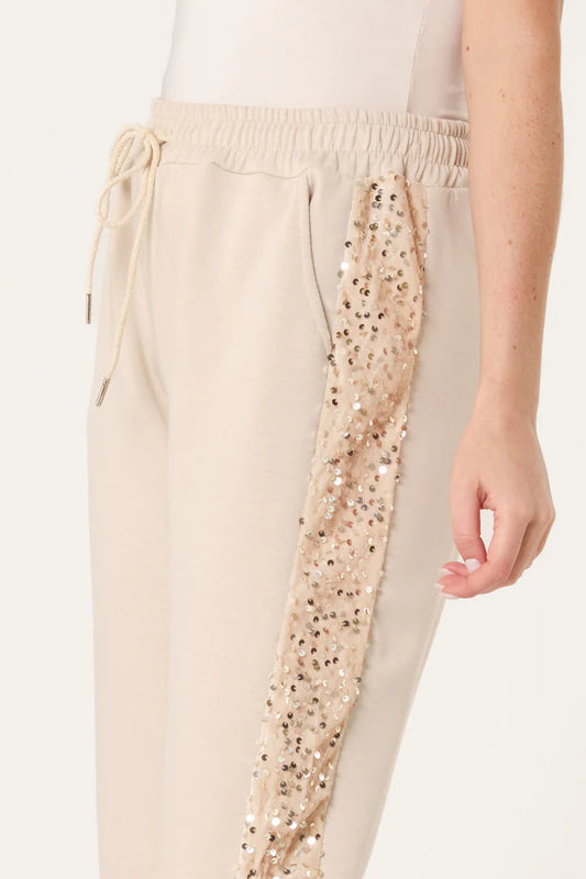 A person wearing cream-coloured sequin velvet joggers featuring an elastic drawstring waist and a beige sequin stripe down the side, ideal for a chic athleisure ensemble.