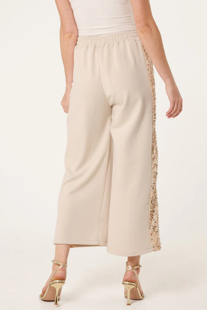 A person wearing wide-leg, cream-coloured sequin velvet joggers from the Italian collection, paired with a white top and strappy heeled sandals, seen from the back.