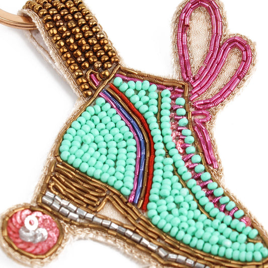 Beaded ornament shaped like a bunny slipper. Features light green, pink, and gold beads with a red ball, showcasing detailed craftsmanship—a delightful handmade treasure for disco enthusiasts and skaters alike.