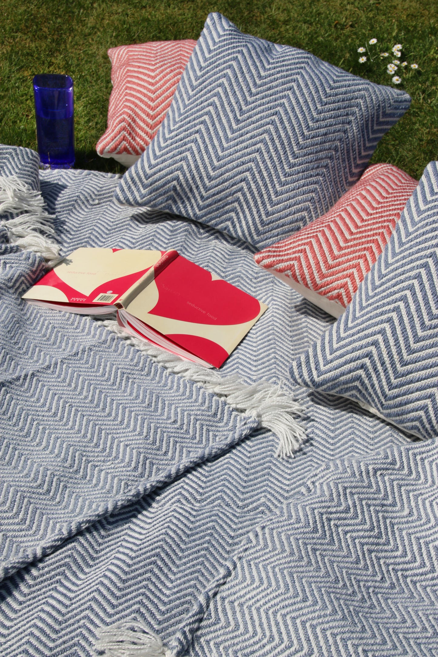 Blue and red chevron-patterned cushions and blankets on grass, alongside an open book and a blue mug, create a cosy nook. Nearby, a sustainable rug crafted from recycled plastic bottles completes the charming outdoor setup.