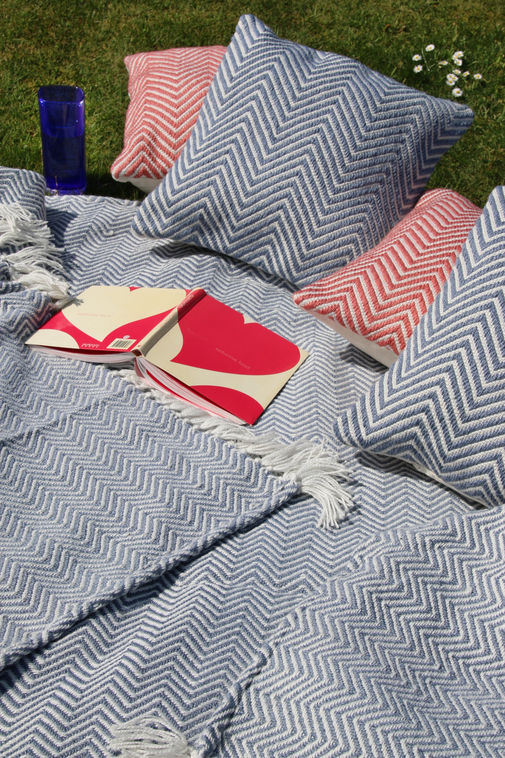 Blue and red chevron-patterned cushions and blankets on grass, alongside an open book and a blue mug, create a cosy nook. Nearby, a sustainable rug crafted from recycled plastic bottles completes the charming outdoor setup.