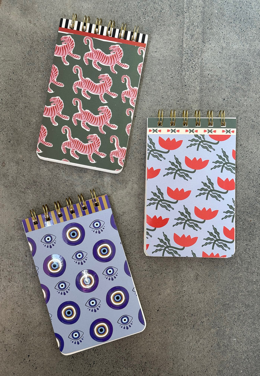 Three spiral-bound lined notebooks with premium paper feature pink tigers, red flowers, and blue evil eye designs, all arranged on a grey surface.