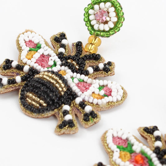 Close-up of a handcrafted, beaded bee-shaped earring with intricate embroidery in black, white, pink, gold, and green beads, attached to a circular floral design centred in pink—a stunning example of beaded bee earrings.