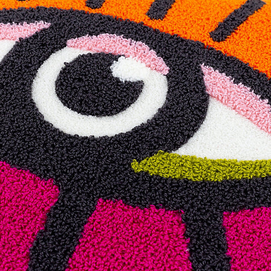 Close-up of a colourful textured rug showcasing a stylised eye design with black, white, pink, orange, and green sections, complemented by a vibrant colourful throw cushion for added flair.