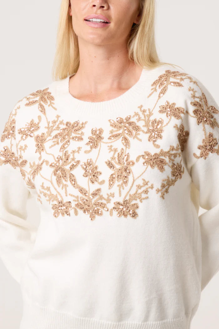 A person wearing a white embroidered floral jumper adorned with gold floral embroidery across the chest and shoulders.