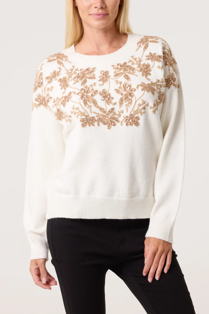 Woman wearing an embroidered floral jumper featuring tan floral designs across the chest, paired with black trousers, standing against a plain background.