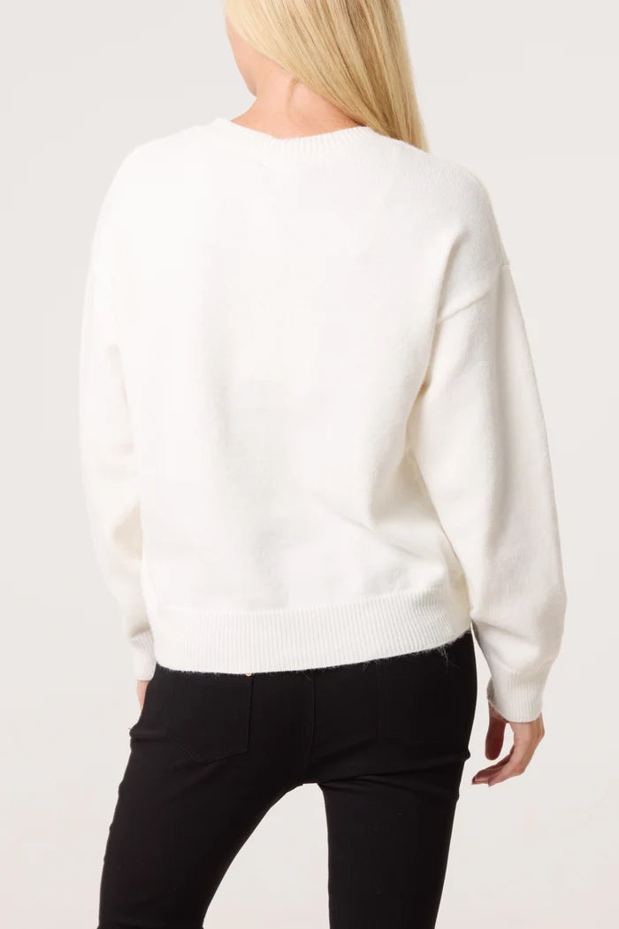 A person with long blond hair wears an embroidered floral jumper and black trousers, facing away from the camera against a plain background.