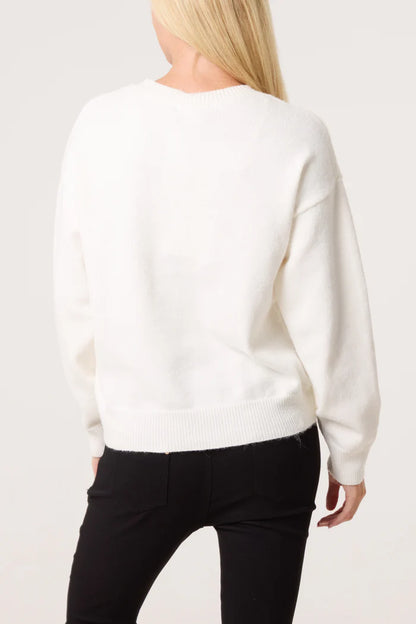 A person with long blond hair wears an embroidered floral jumper and black trousers, facing away from the camera against a plain background.