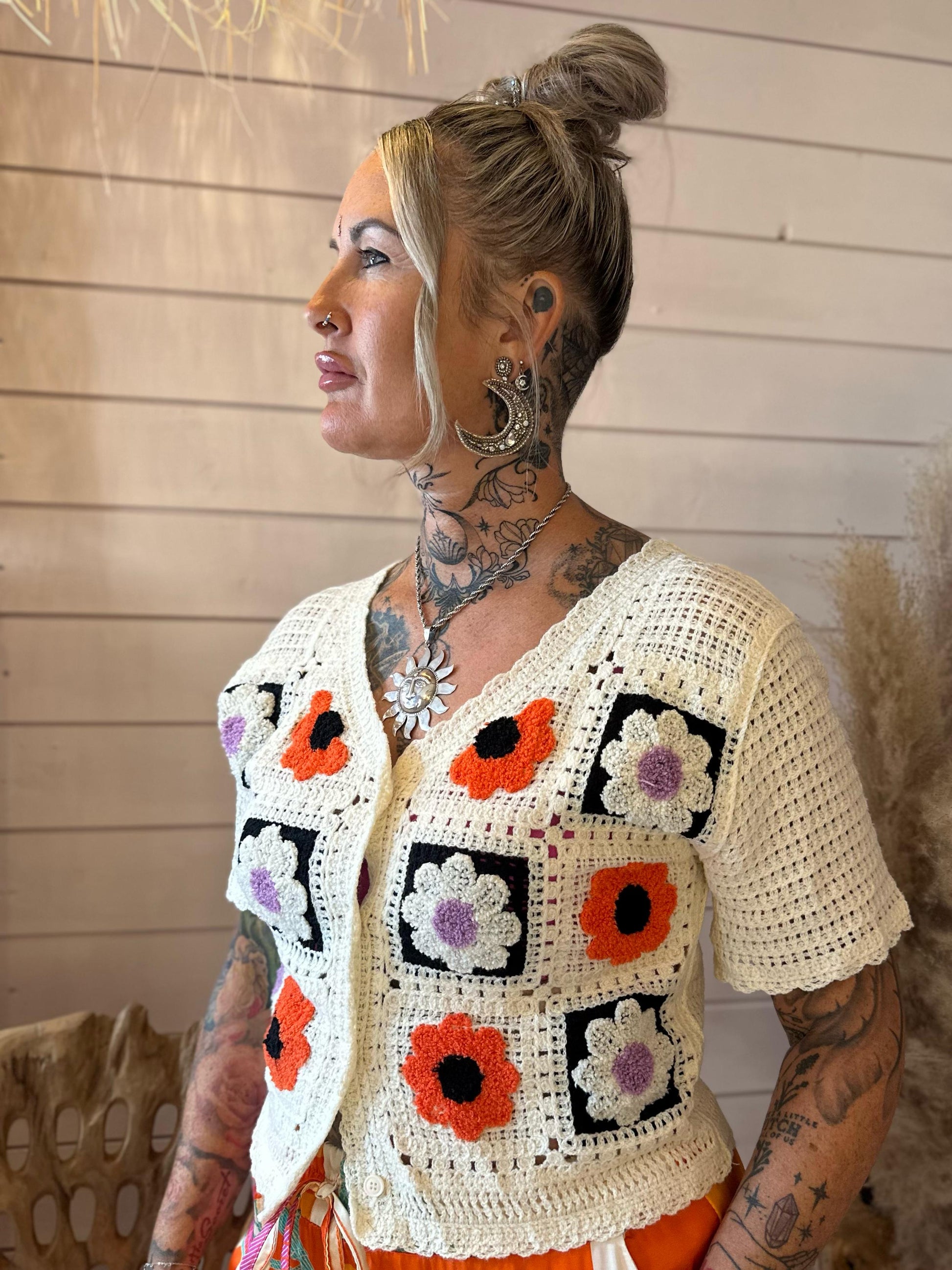 A woman with tattoos and a nose ring is wearing a luxurious, short-sleeved crochet top featuring colourful floral squares, standing indoors against a wooden wall.