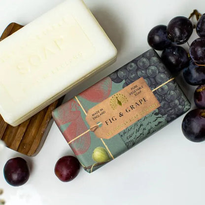 Two bars of vegan-friendly Anniversary Fig and Grape Soap, one unwrapped and one in decorative packaging, shown next to fresh grapes and a wooden soap dish.