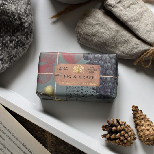 An Anniversary Fig and Grape Soap bar, suitable for vegans and wrapped in decorative paper and twine, sits on a table with pine cones, linens, and an open book close by.