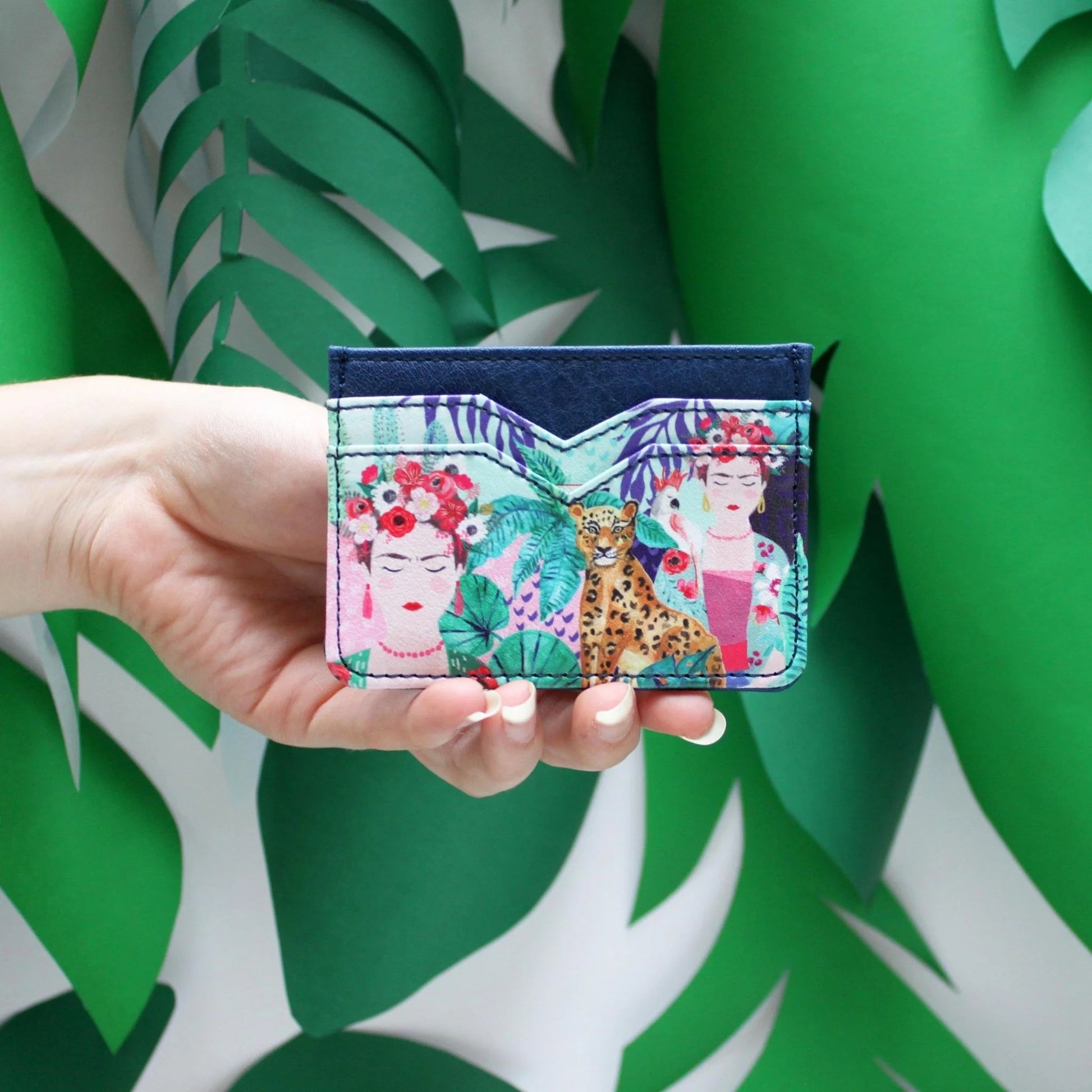 A hand holds a vibrant card holder with a tropical print, displaying illustrations of two women wearing floral headpieces and a cheetah, all set against lush green foliage.